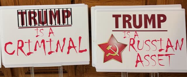 Two white protest signs side-by-side. The left sign has 'TRUMP IS A CRIMINAL' in red handwritten text with a black bar over…