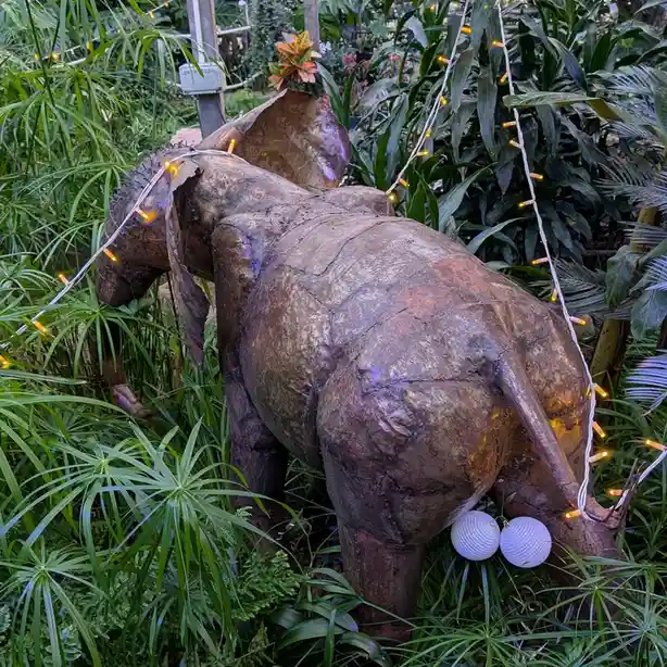 A large, rustic elephant statue made of patched, brown material stands in a lush green garden, adorned with string lights…