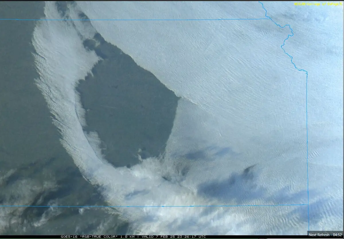 A satellite image from GOES-16 shows a large, dark, roughly circular landmass surrounded by white clouds and snow, with…