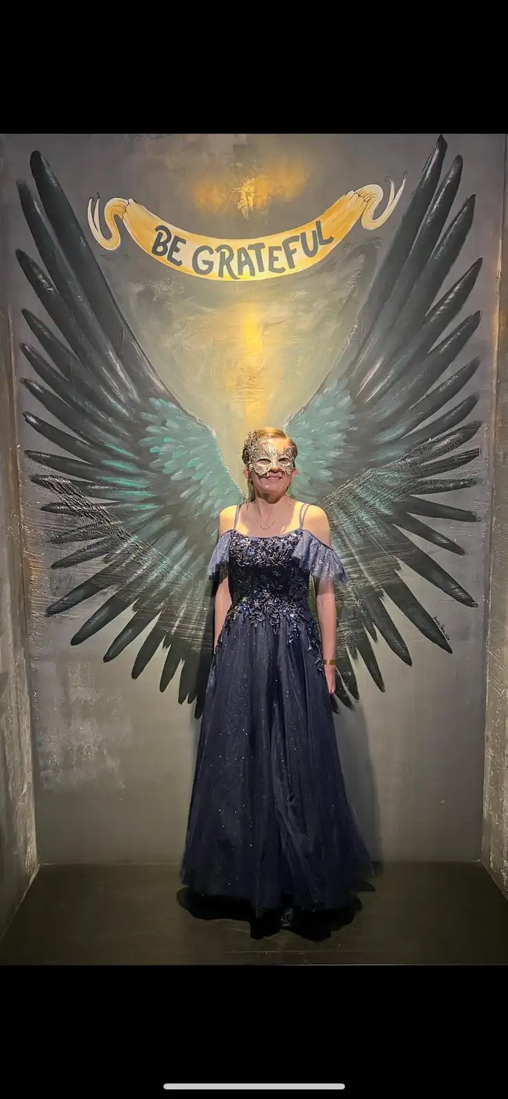 A person wearing a dark blue, sparkly gown and an ornate mask stands in front of a painted backdrop depicting large, dark…