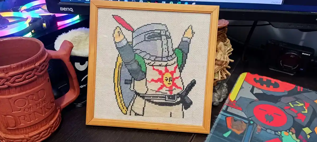 A framed cross-stitch artwork depicting a pixelated, armored character holding a mug with a sun design, placed on a desk…