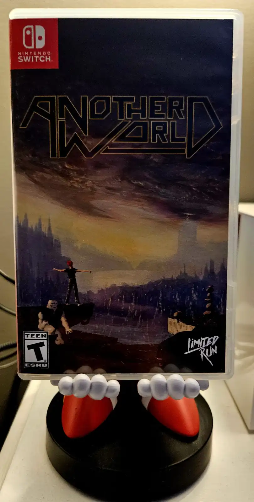 A Nintendo Switch game case for 'Another World' by Limited Run Games is displayed on a black stand shaped like a pair of…