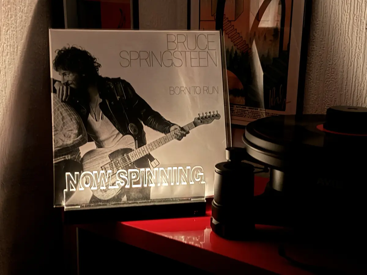 A black-and-white album cover of Bruce Springsteen's 'Born to Run' is displayed upright on a red shelf next to a turntable…