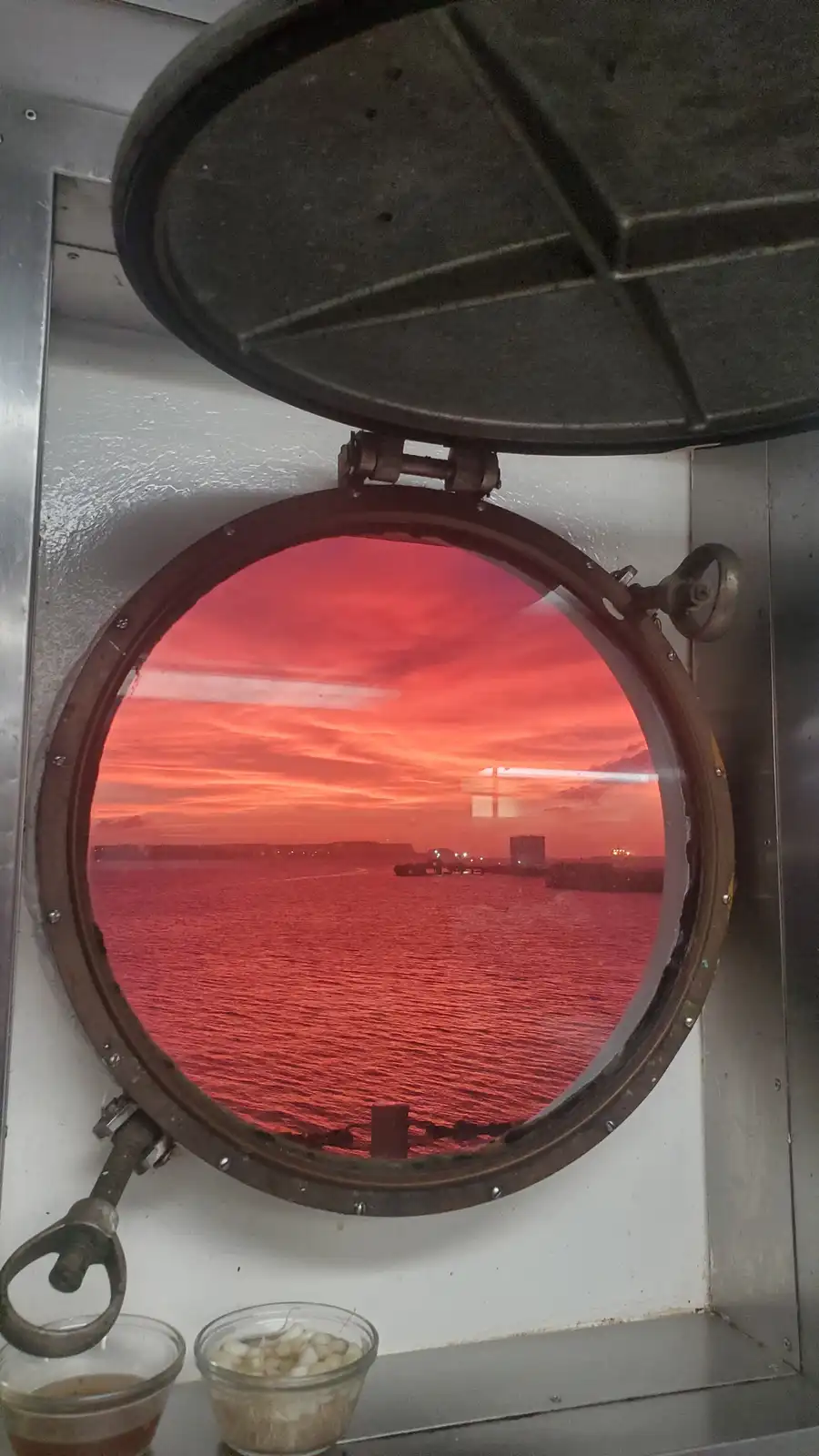 A view through an open, circular porthole on a ship or boat shows a stunning red-orange sunset over water, with distant land…
