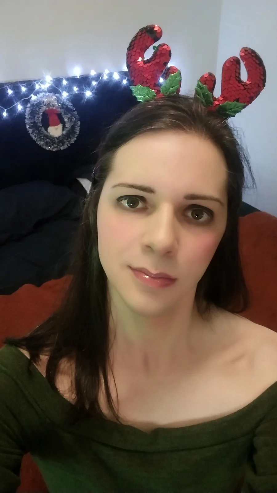 A woman with dark hair and a green top wears red sequined reindeer antlers with green holly leaves. Behind her, a dark…