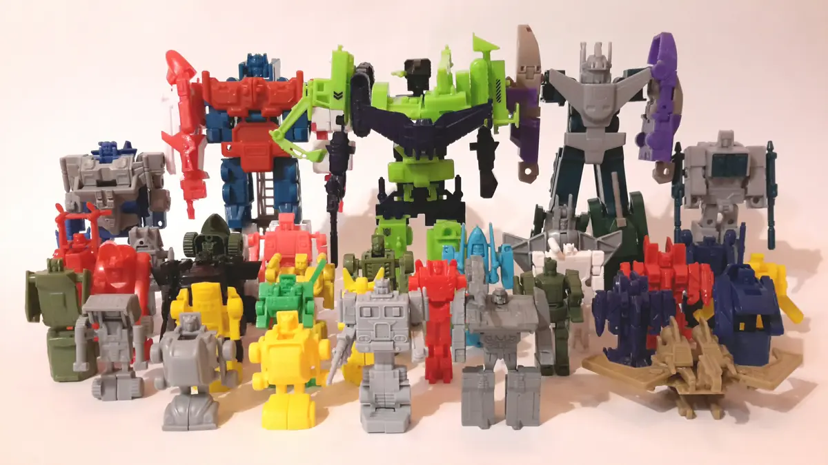 A large collection of colorful Transformers action figures is arranged in rows on a plain white surface, showcasing various…