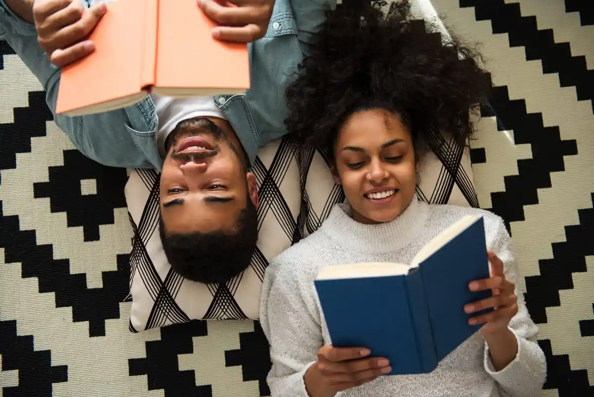 Two people lie on patterned pillows on a black-and-white geometric rug, each holding an open book. The person on the left…