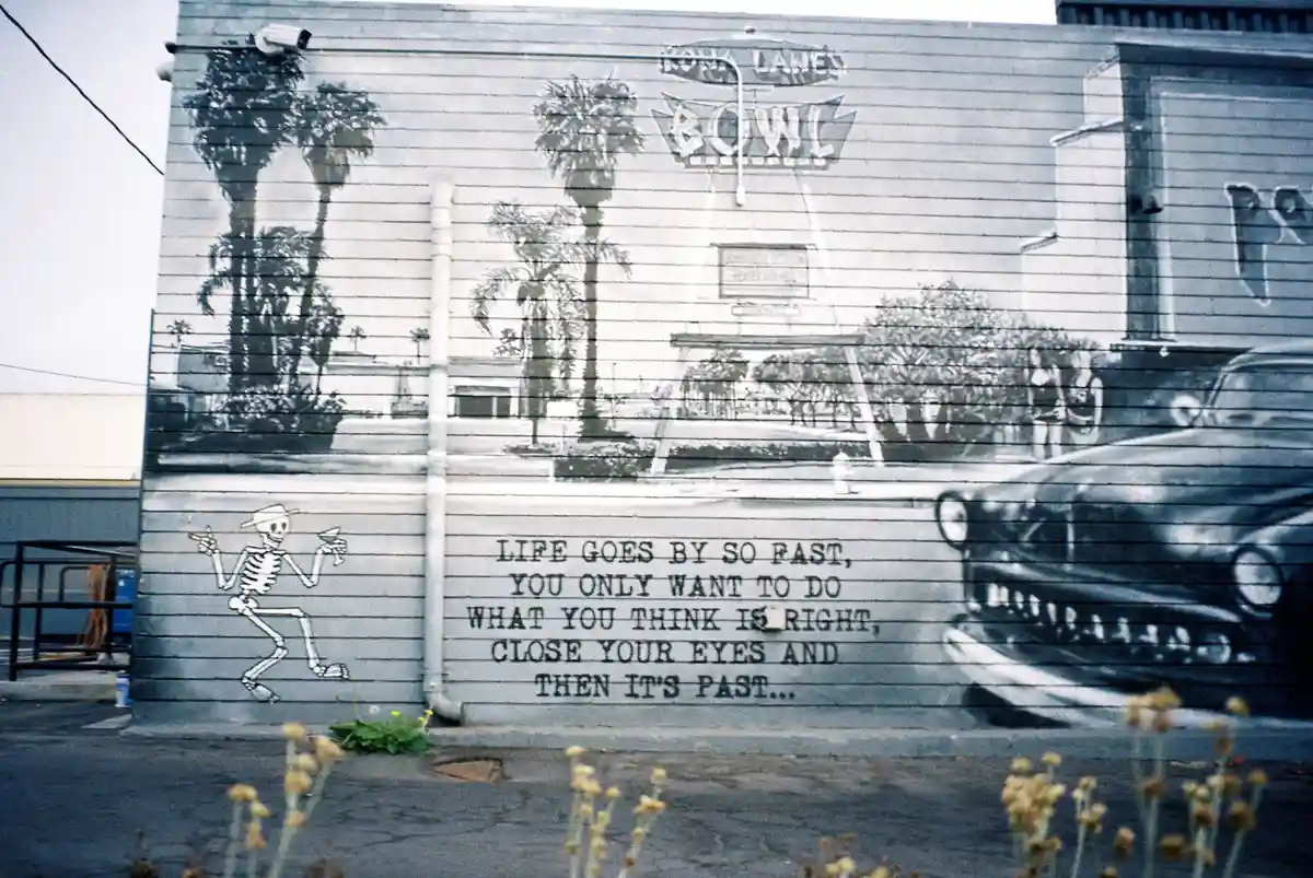 A black-and-white mural on a building depicts a vintage bowling alley called 'Rona Lane Bowl' with palm trees and a classic…
