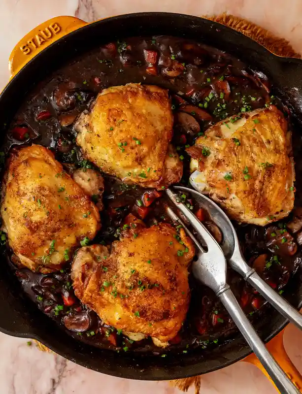 A top-down photo of four golden-brown chicken thighs in a dark, rich sauce with mushrooms and chopped chives, served in a…