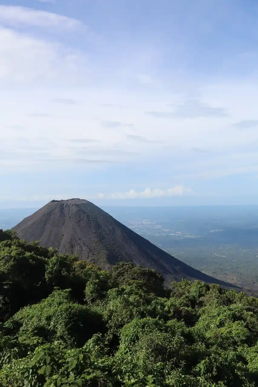 A scenic photo shows a large, dark volcanic cone rising above a lush green forest foreground, with a hazy valley and distant…