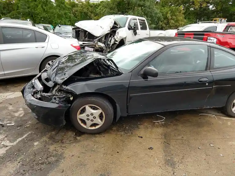A black sedan with a severely damaged front end, hood crumpled and engine exposed, sits in a junkyard surrounded by other…