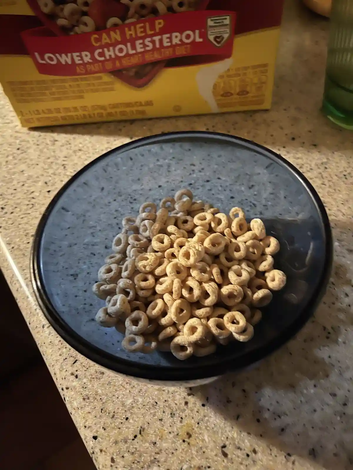 A bowl of plain cereal rings sits on a speckled countertop, with a cereal box behind it that prominently displays the text…