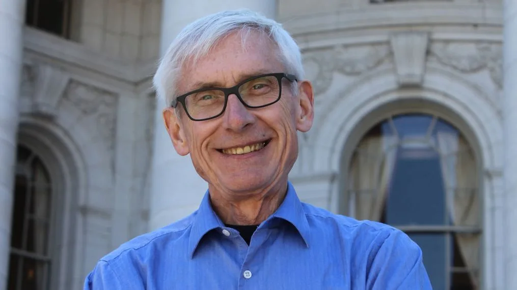 a smiling older man with white hair and black-rimmed glasses wears a blue button-down shirt, standing in front of a…