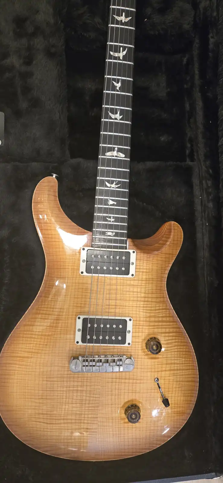 A light-wood electric guitar with a glossy finish and bird-shaped inlays on its black fretboard rests in a black plush case…