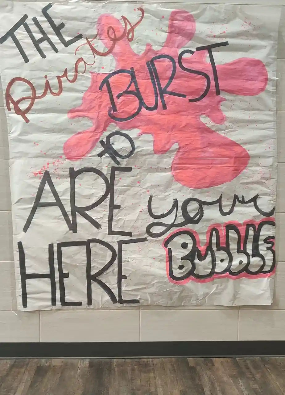A handmade poster on crumpled white paper features large, hand-drawn text in black marker with pink paint splatters. The…