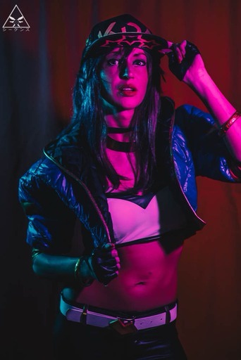 A woman in a shiny blue jacket, black crop top, and a patterned cap poses under dramatic red and blue lighting. She holds…
