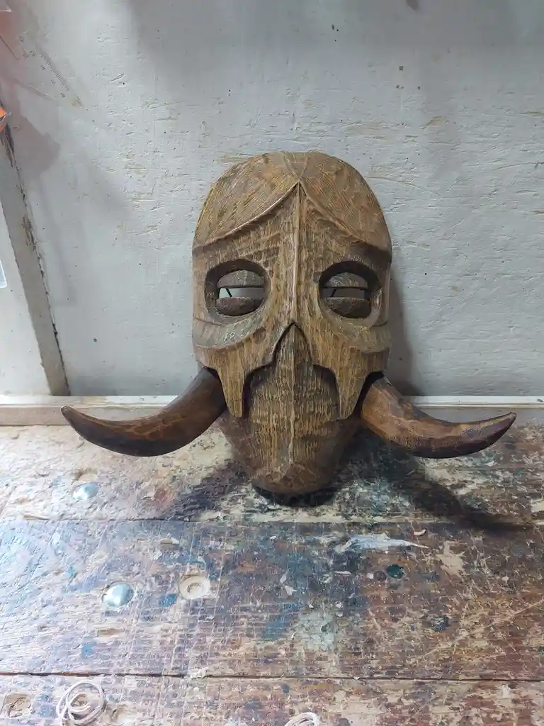 A hand-carved wooden mask with two curved horns protruding from the sides sits on a worn, paint-splattered workbench against…