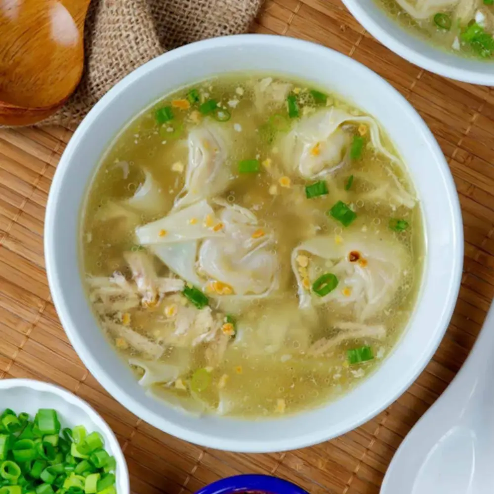 A top-down photo of a white bowl filled with wonton soup, garnished with chopped green onions and minced garlic, resting on…