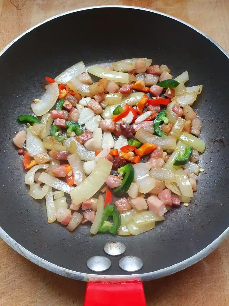 A close-up photo of a black frying pan with a red handle, sitting on a wooden surface, filled with sautéed chopped onions…