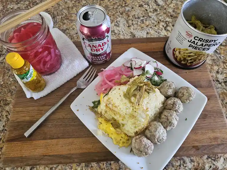 A plate of food including rice, eggs, meatballs, pickled onions, radishes, and crispy jalapeño pieces sits on a wooden…