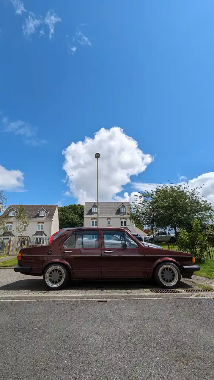 A maroon vintage sedan is parked on a paved street under a bright blue sky with a large, fluffy white cloud directly above…