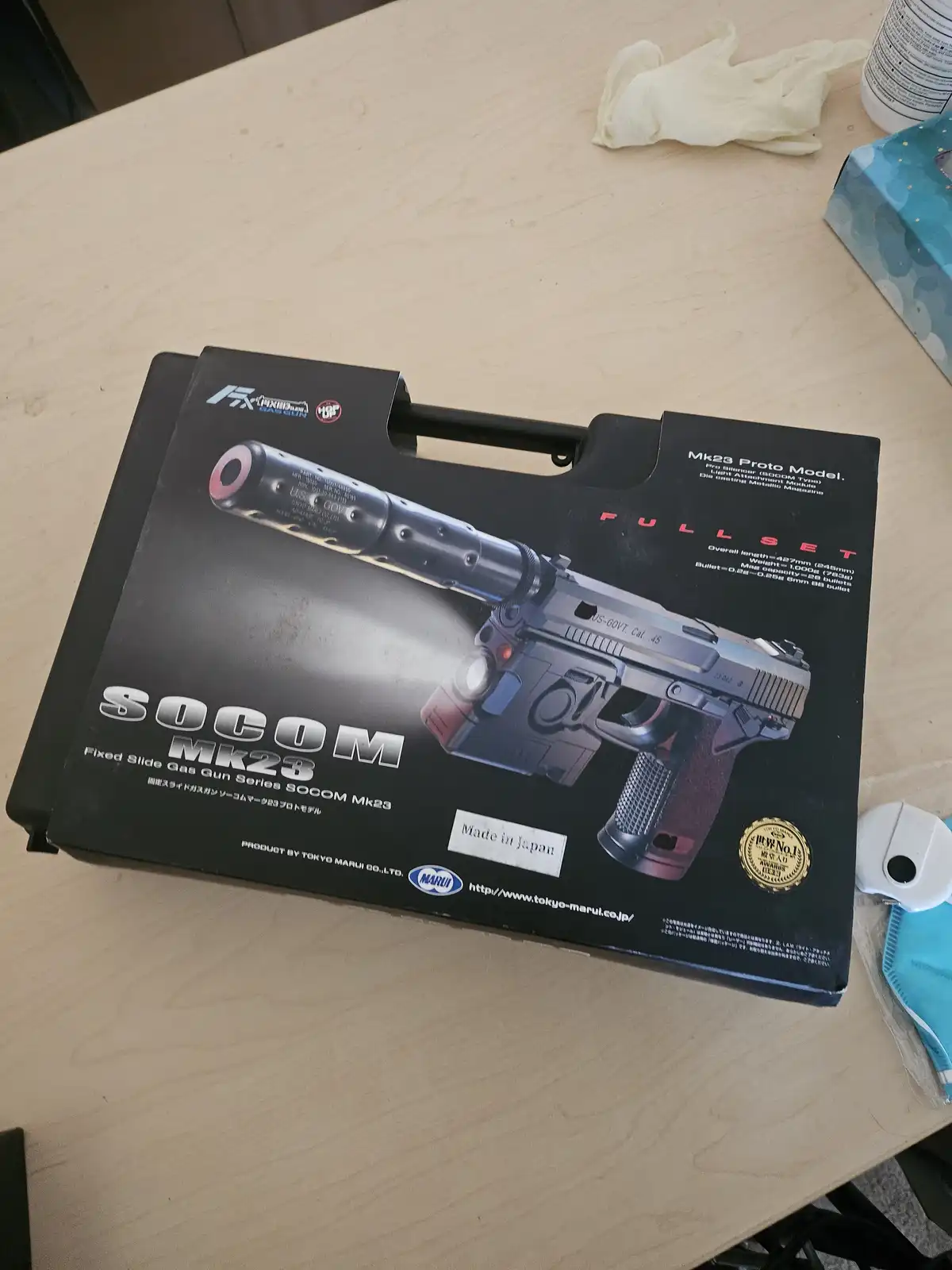 A black box for a Tokyo Marui SOCOM Mk23 gas gun sits on a wooden table, featuring an image of the silver and black pistol…