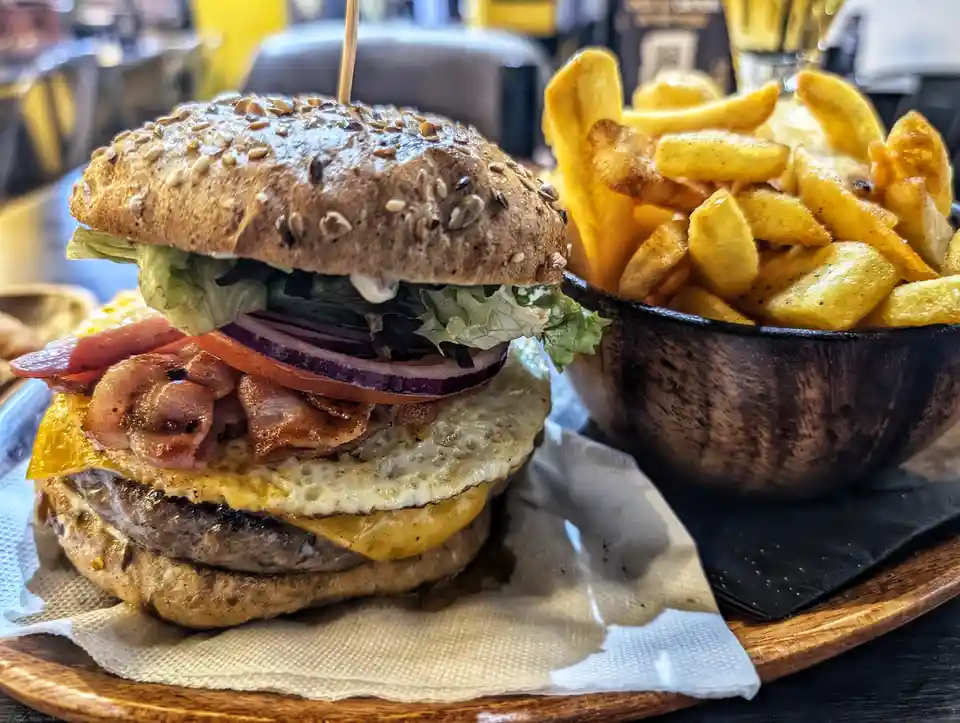 A close-up photo of a large, layered burger with a sesame seed bun, bacon, egg, cheese, tomato, onion, and lettuce, served…