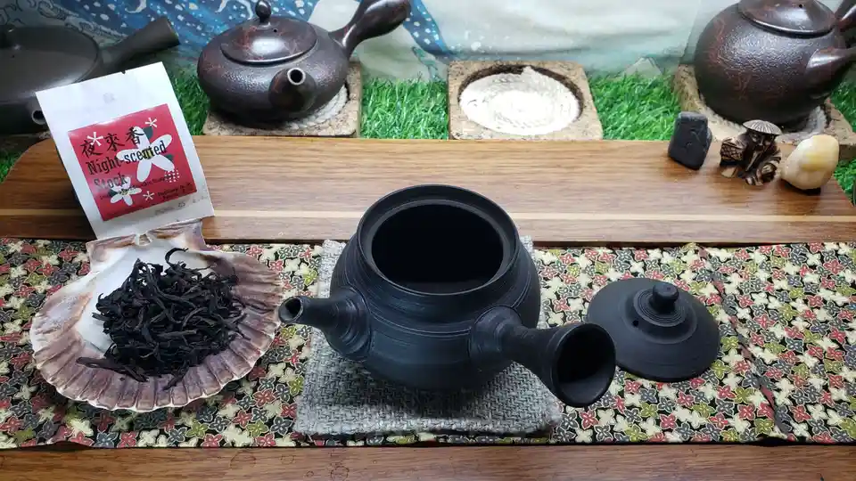 A black ceramic teapot with its lid off sits on a patterned cloth, next to a shell holding dark tea leaves and a bag labeled…