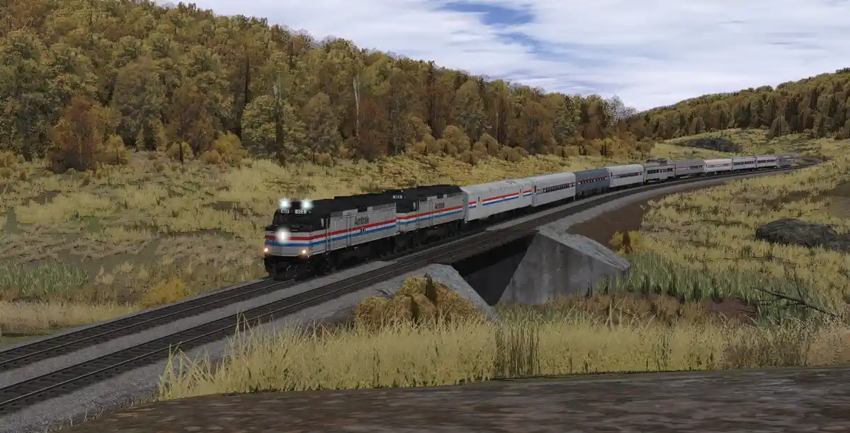 An Amtrak passenger train with locomotives and silver cars travels across a concrete bridge through a grassy, forested…