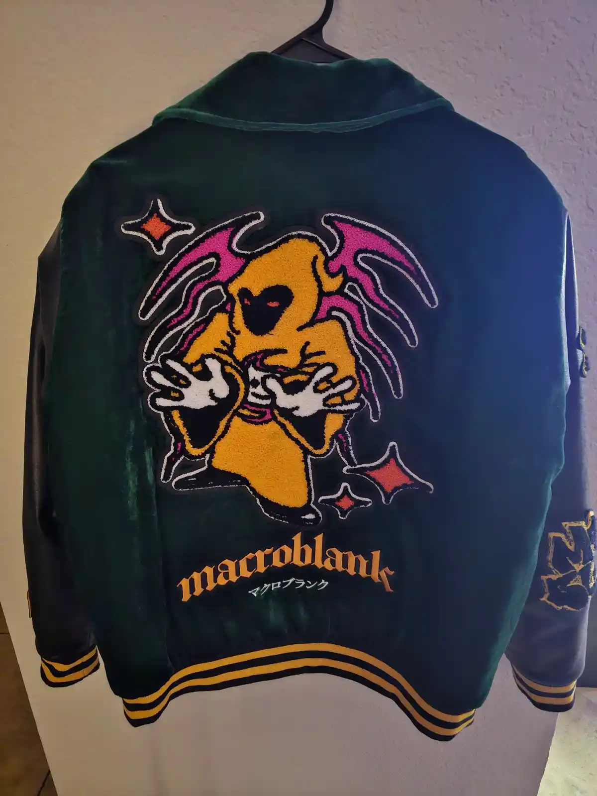 A dark green velvet varsity jacket with black leather sleeves and yellow trim hangs on a hanger. The back features a large…