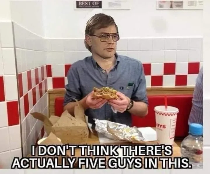 A man with glasses sits in a diner booth holding a partially eaten sandwich, looking confused. On the table are a cardboard…