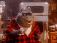 A still from the movie 'The Muppet Movie' shows Kermit the Frog wearing a red plaid shirt and holding a glass. He is seated…