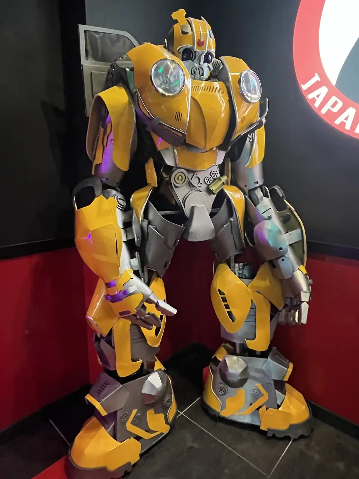 A large, yellow and silver robot statue resembling Bumblebee from Transformers stands in a corner with red and black walls…