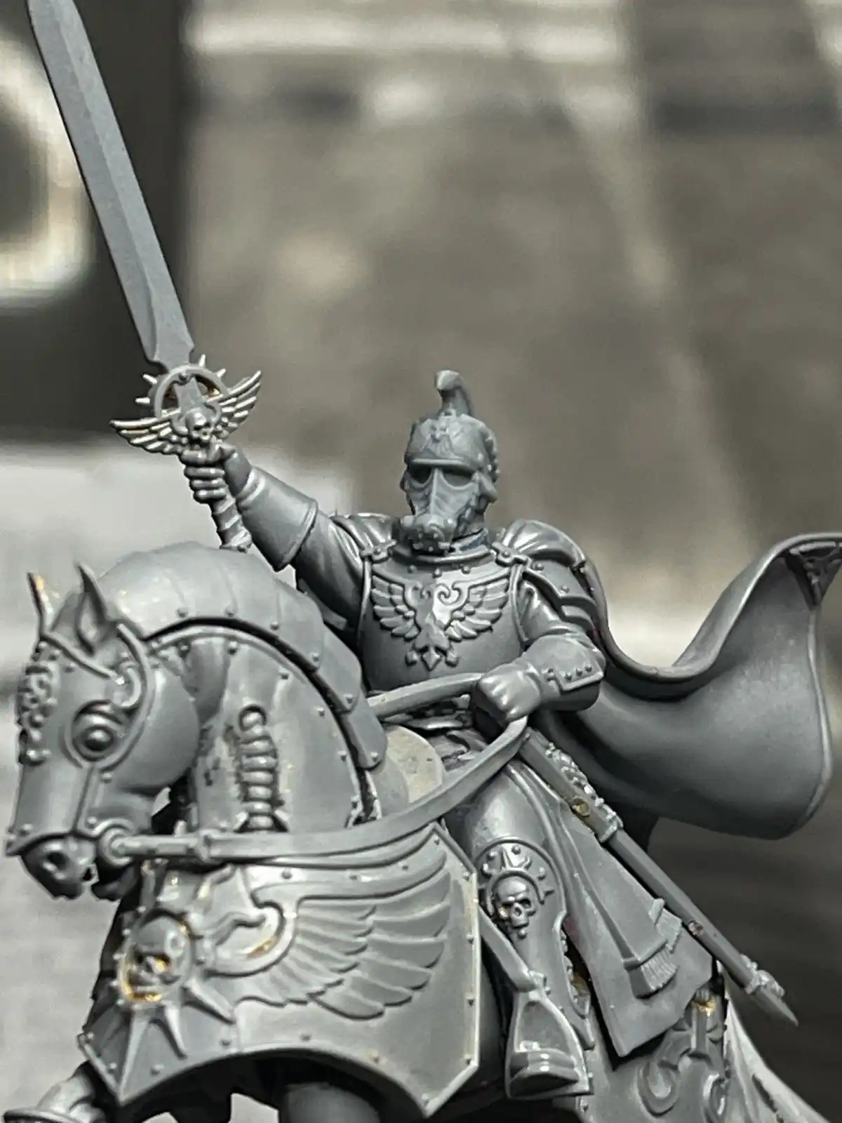 A close-up of a grey-painted miniature of a heavily armored knight on a similarly armored horse, holding a sword aloft. The…