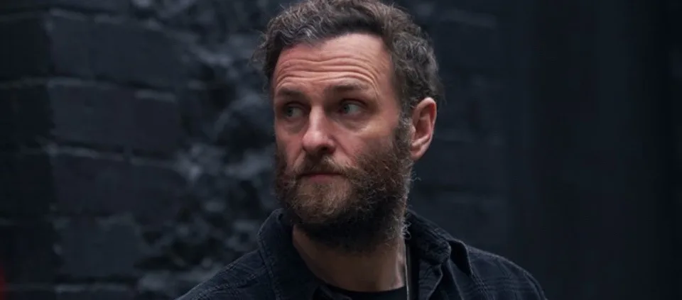 a man with a beard and dark hair looks sideways with a skeptical expression, standing in front of a dark brick wall. The…