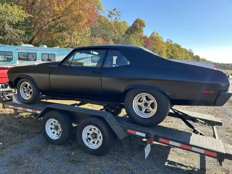 A matte black classic muscle car, likely a Chevrolet Chevelle, is parked on a flatbed trailer in an outdoor lot with autumn…