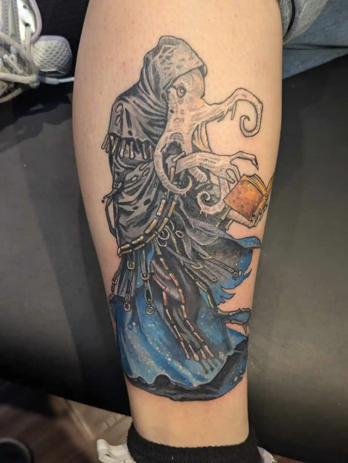 A tattoo on a person's lower leg depicts a hooded, octopus-like creature with multiple tentacles, holding an open book. The…