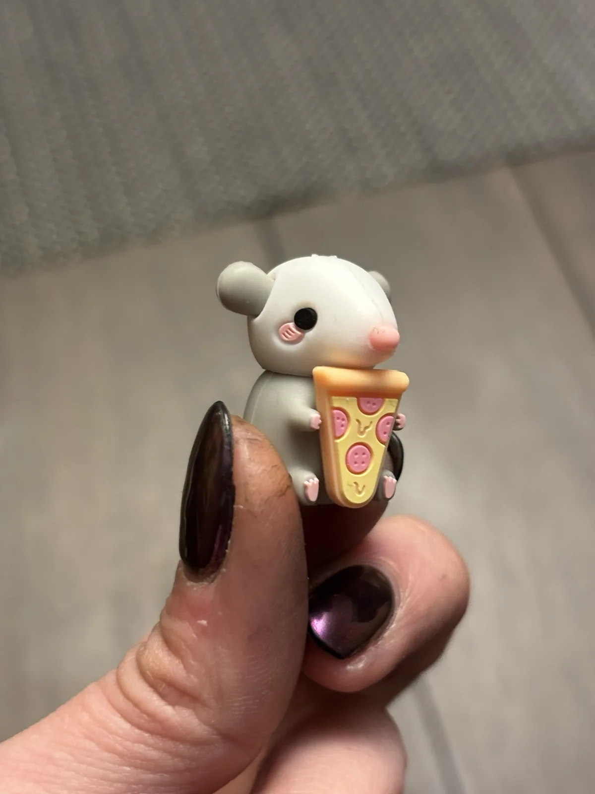 A small, white, cartoonish mouse figurine with pink cheeks and nose is being held between a person's thumb and forefinger…