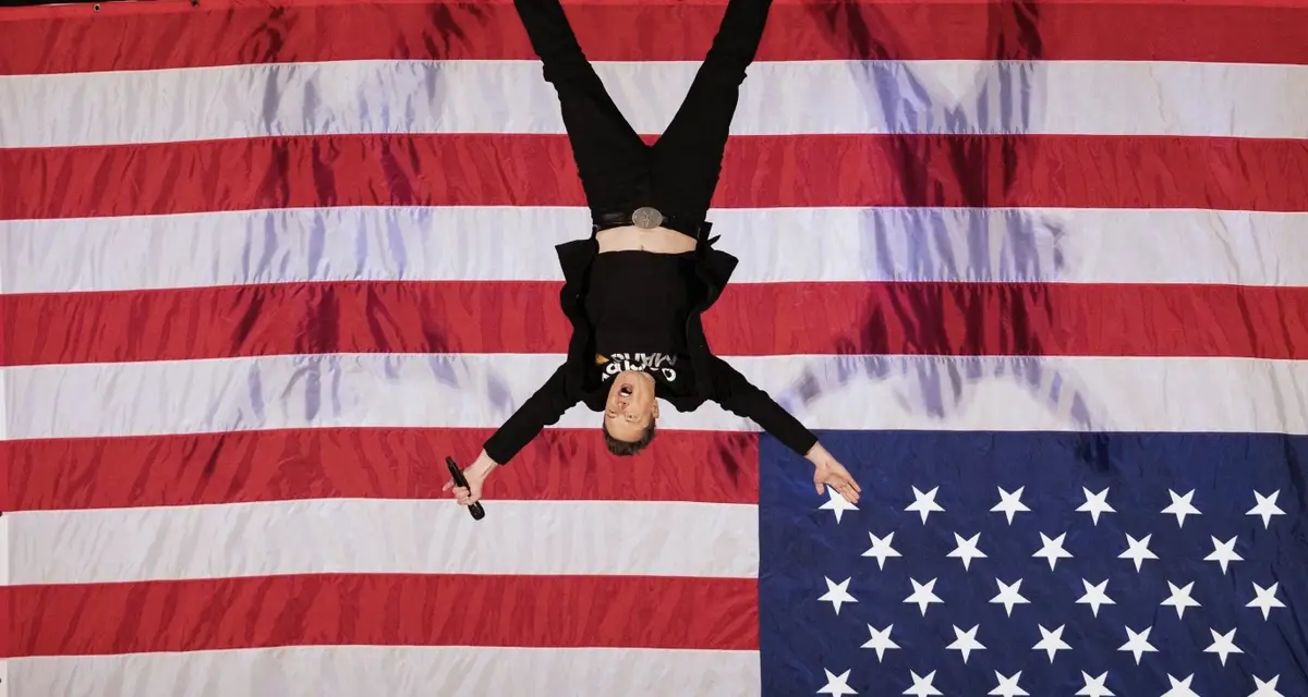 A man wearing a black shirt and pants, holding a microphone, is lying upside down on a large American flag. His head is near…