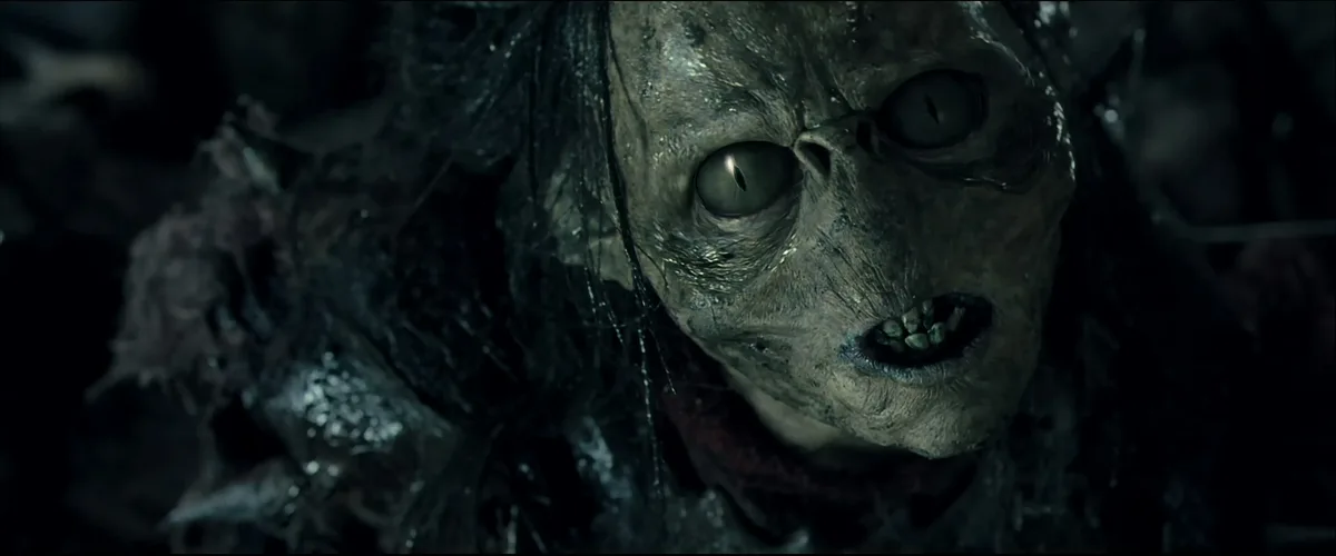A close-up shot of a grotesque, decaying humanoid creature with pale, wrinkled skin, large cat-like eyes, and visible teeth…