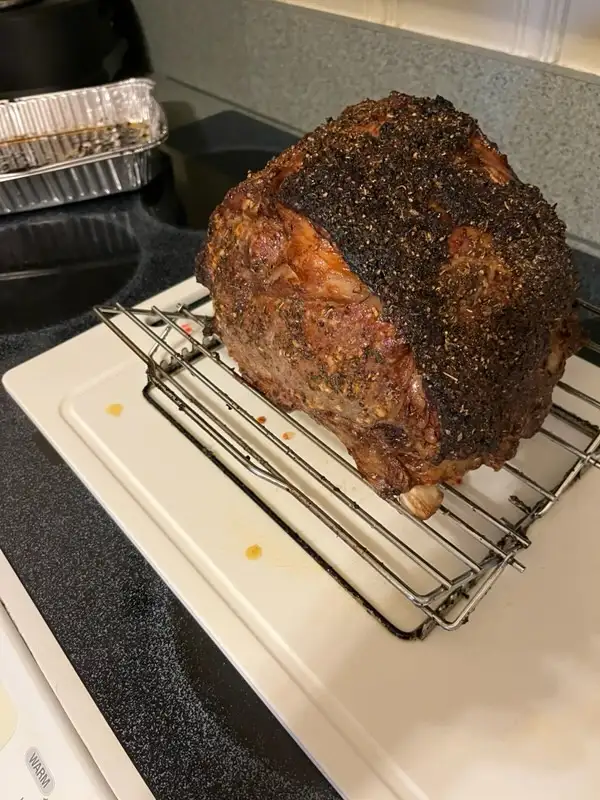 A large, roasted meat joint with a dark, seasoned crust sits on a metal cooling rack atop a white cutting board on a kitchen…