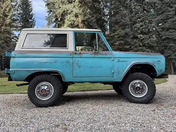 A vintage light blue Ford Bronco with a white roof and visible rust patches is parked on gravel, featuring large Mickey…