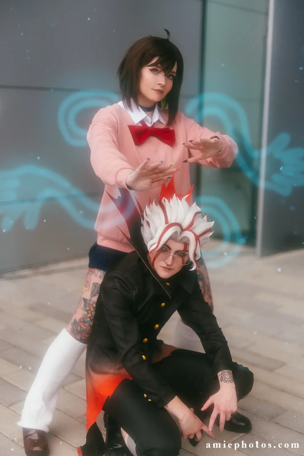 Two cosplayers pose outdoors: one crouches in a black outfit with white and red spiky hair and glasses, while the other…