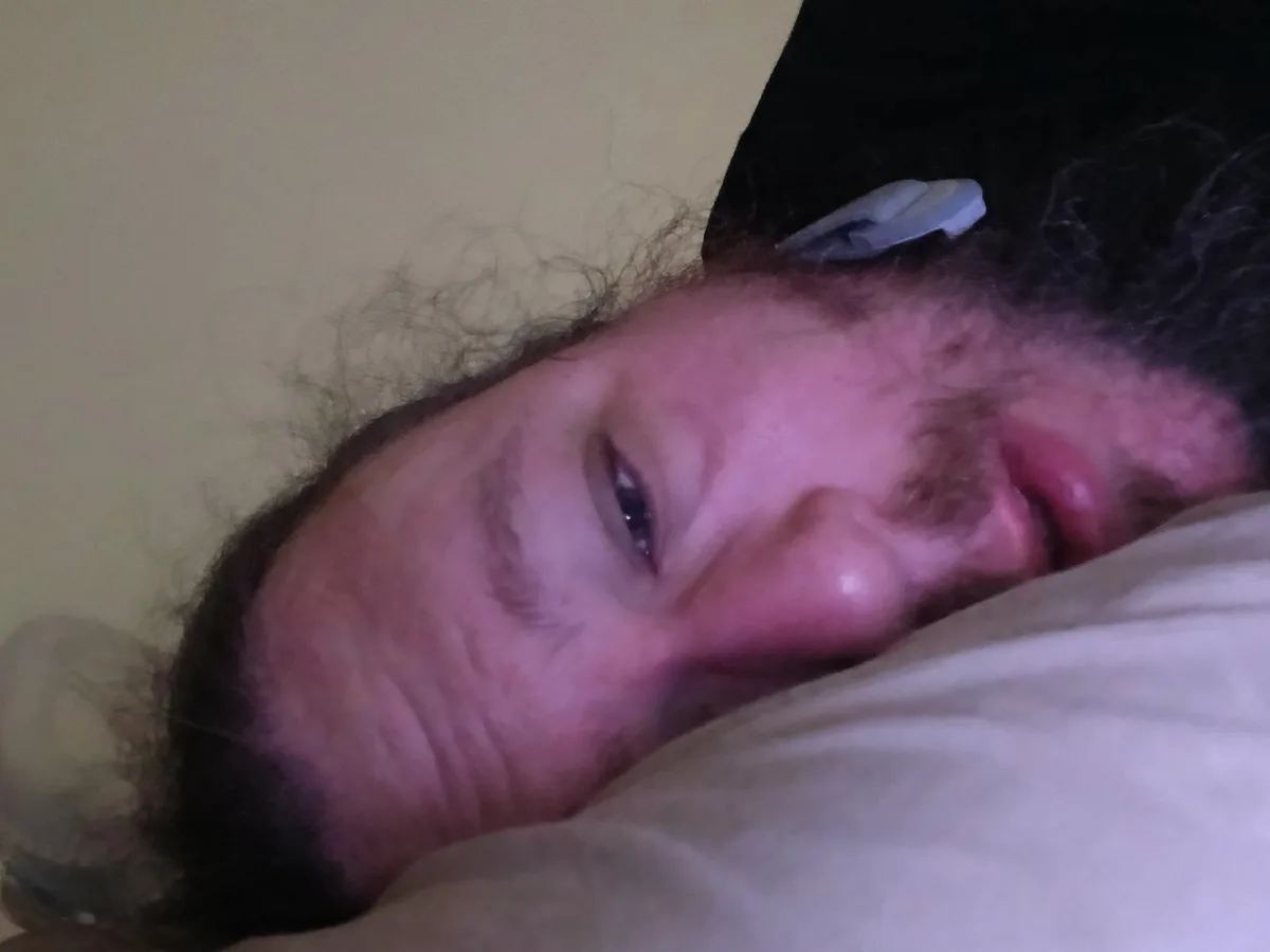 A person with dark curly hair and a beard is lying in bed, half-asleep with one eye open, looking at the camera. A small…