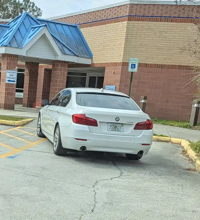 A white BMW 550i is parked in a designated handicapped parking spot at a government building, directly under a sign that…