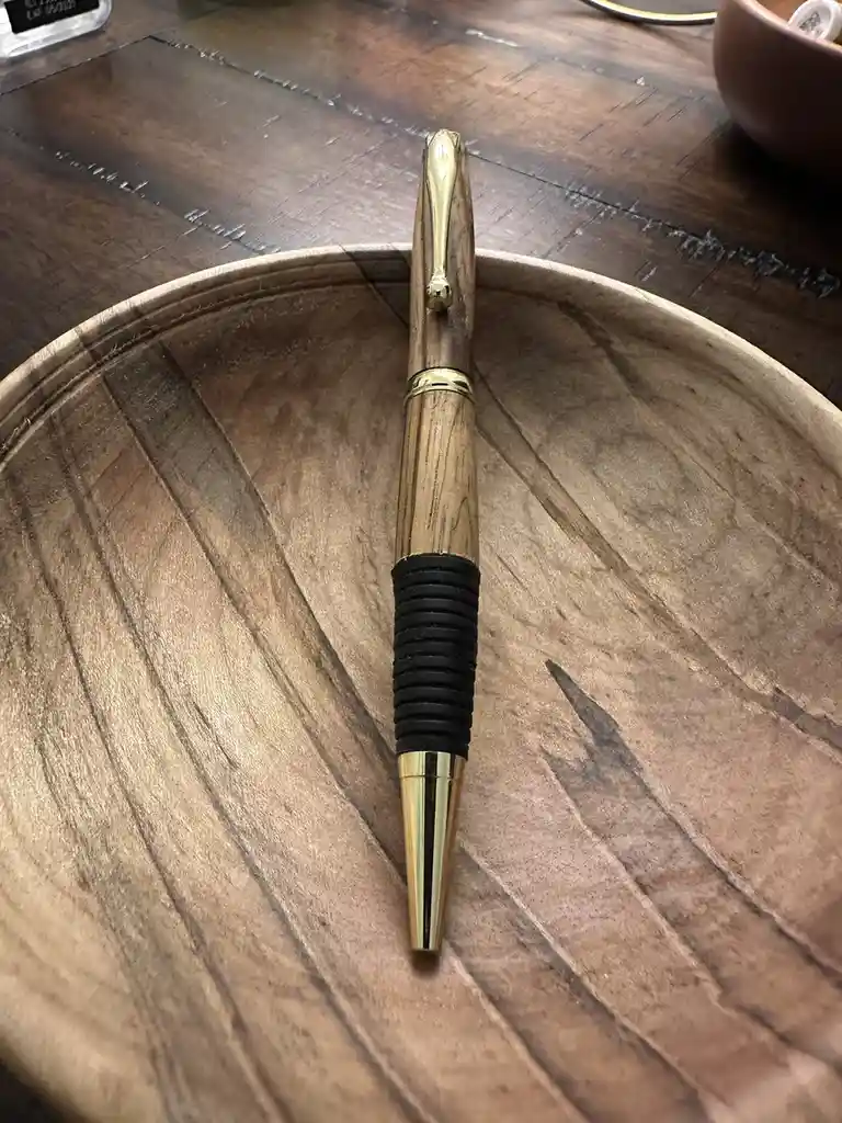 A close-up photo shows a decorative pen with a wooden body, gold-colored metal accents, and a black rubber grip, resting…
