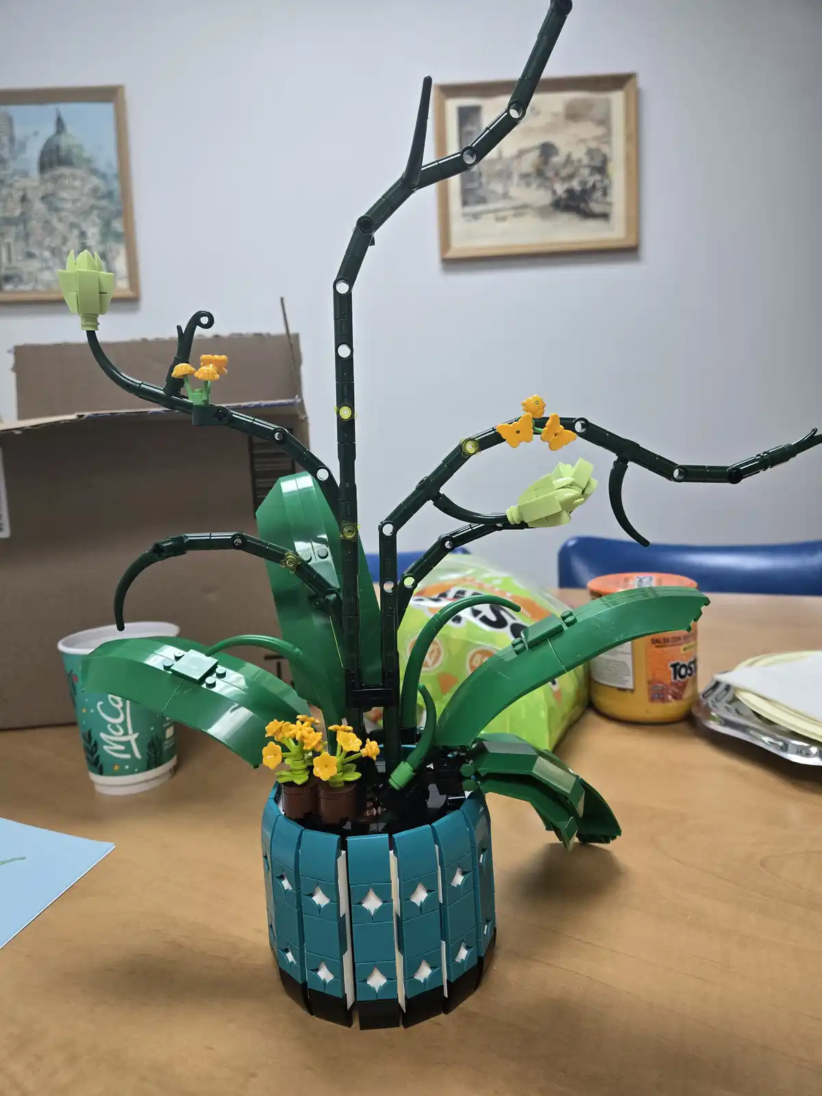 A detailed LEGO model of a potted plant with green stems, large leaves, and yellow flowers sits on a wooden table. The plant…
