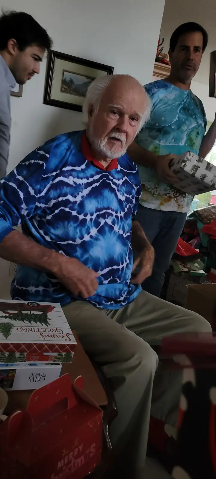 An older man with white hair and a beard, wearing a blue tie-dye shirt, sits surrounded by Christmas gifts and looks…