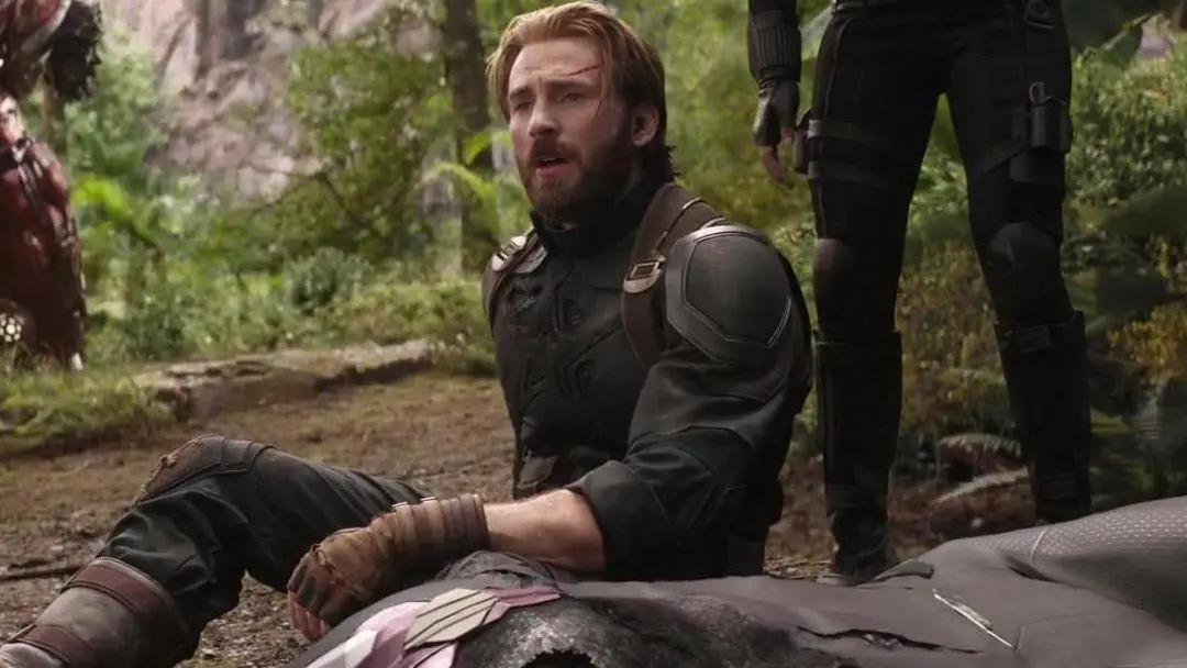 A man with a beard and a cut on his forehead sits on the ground in a forest, looking shocked and confused, with another…