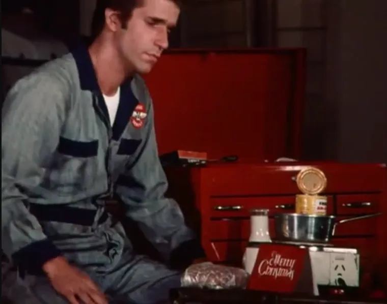 a man in a grey work jumpsuit sits dejectedly beside a red toolbox, looking down at a wrapped gift. On the toolbox, a pot…